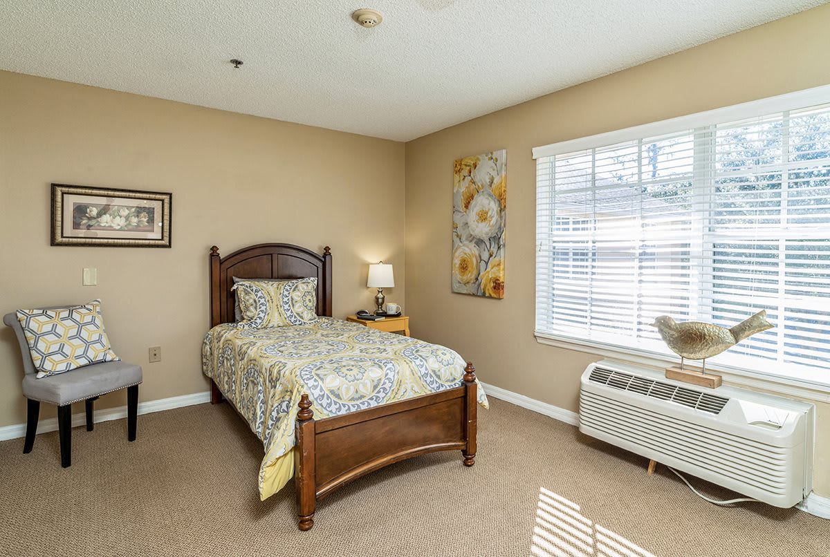 A cozy bedroom with a single bed, decorated with floral bedding and artwork, featuring a bedside lamp and a window with blinds.