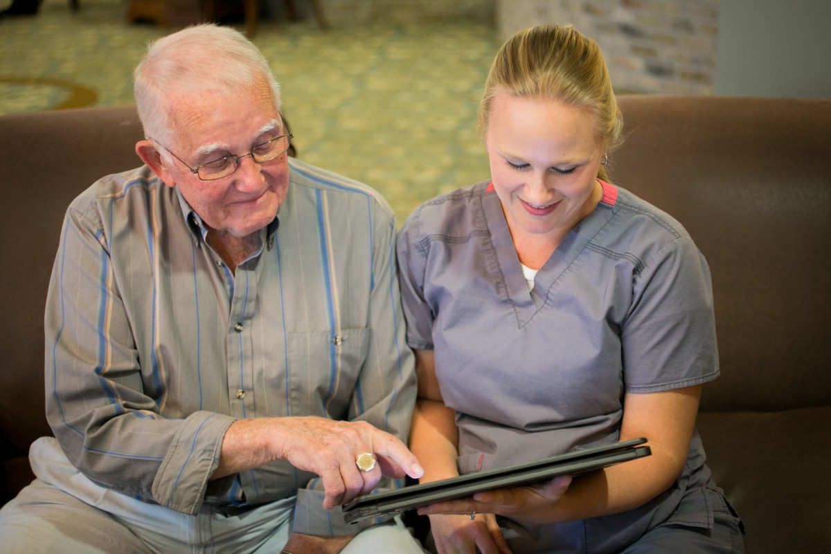 An elderly man and a nurse smiling while looking at a tablet together.