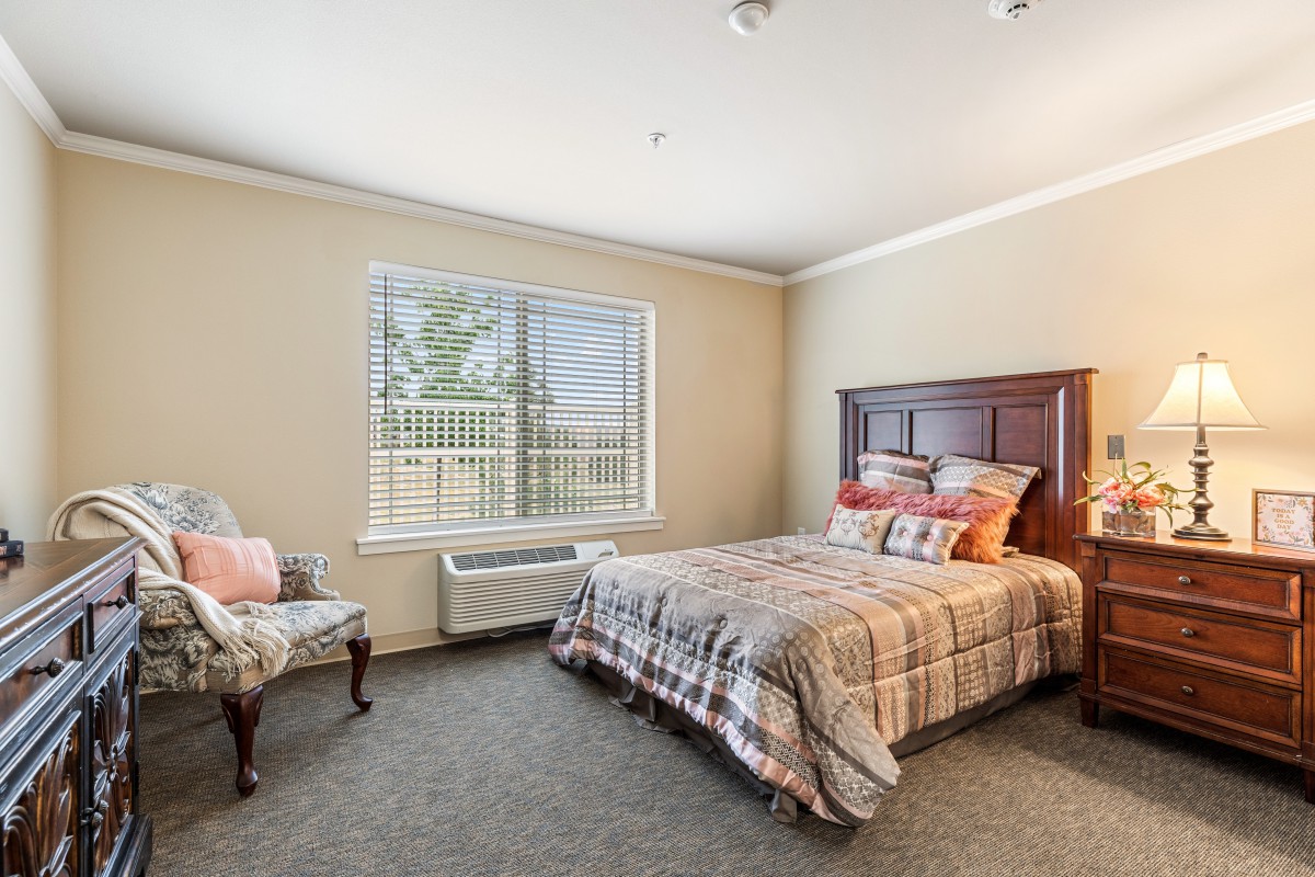 A cozy bedroom with a neatly made bed, an armchair, and a large window providing natural light.