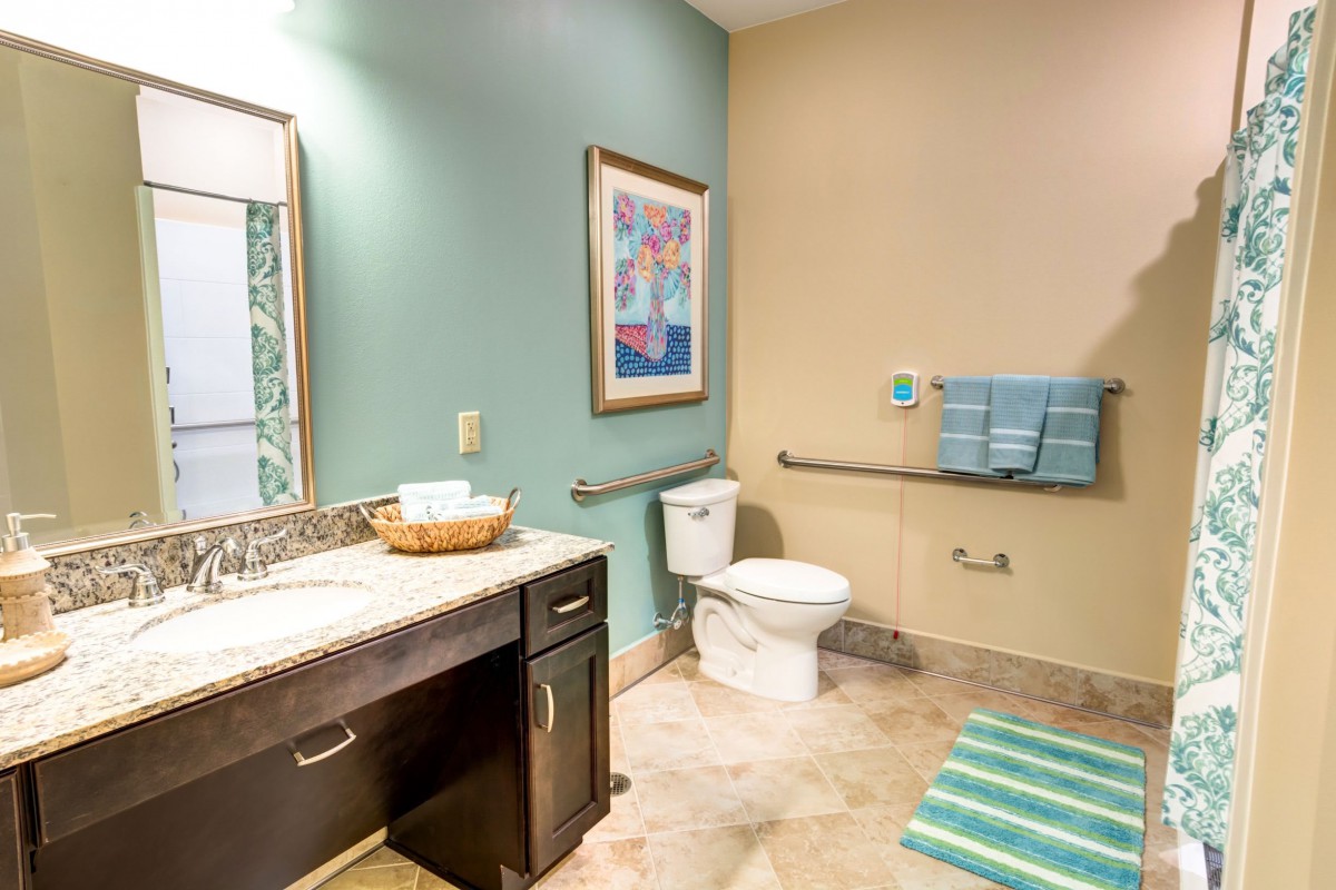 A clean and modern bathroom with teal and beige walls, featuring a toilet, sink, mirror, and colorful artwork.