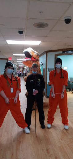 A playful Halloween scene in a hospital with two costumed prisoners, a police officer, and a large dinosaur inflatable.