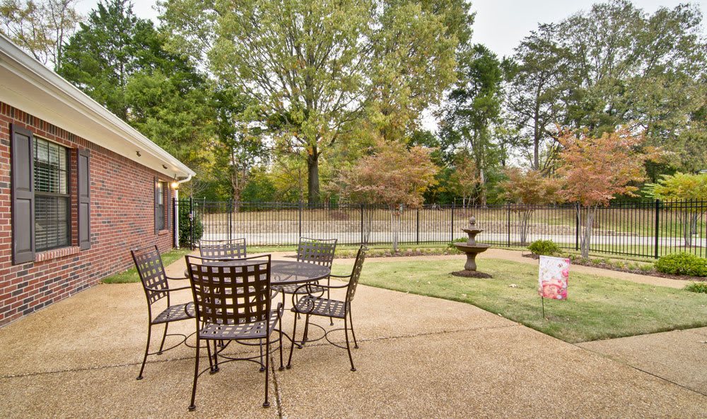 A tranquil patio area with a table and chairs overlooks a landscaped garden featuring a fountain and surrounded by trees.