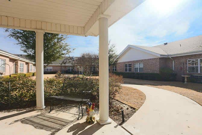 A sunny day view of a porch with a bench, overlooking a pathway and residential buildings.