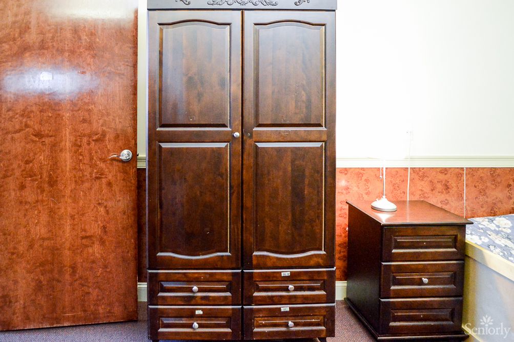 A wooden wardrobe with intricate carvings stands next to a matching drawer unit in a room.