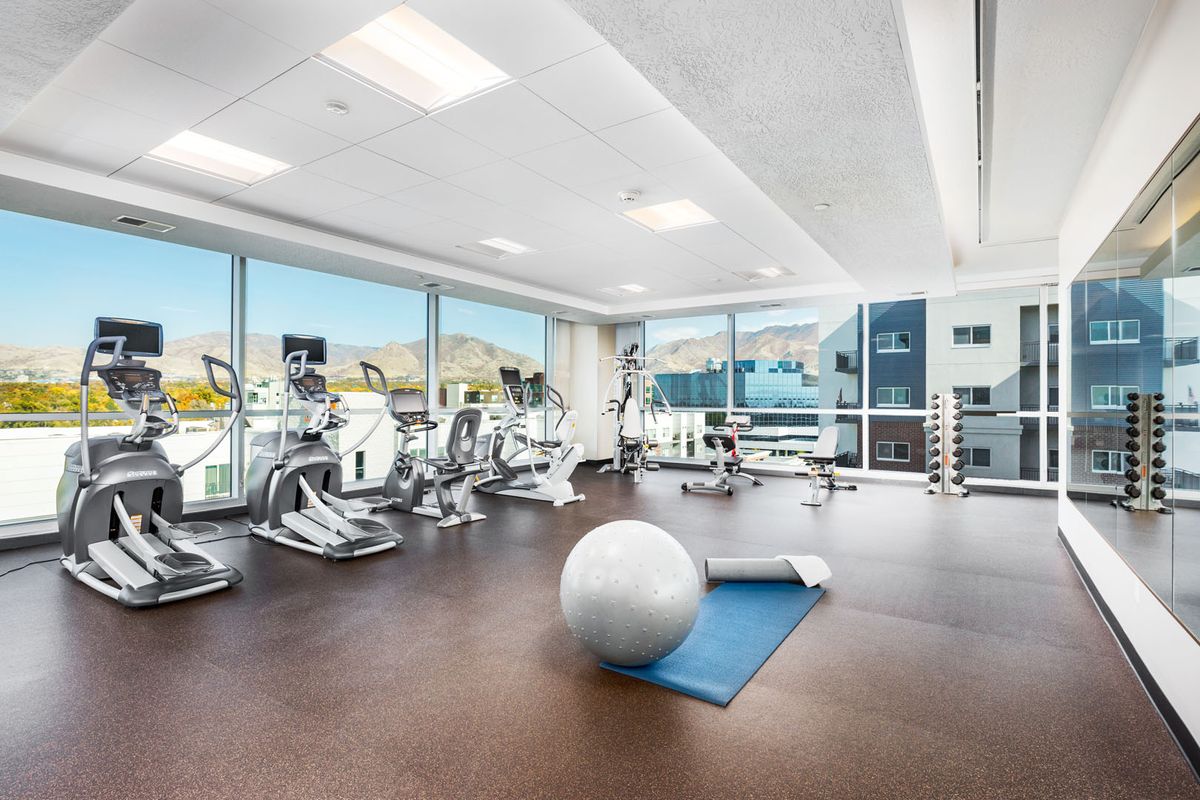 A modern gym featuring a variety of exercise equipment, including treadmills and ellipticals, with a yoga mat and fitness ball in the foreground.