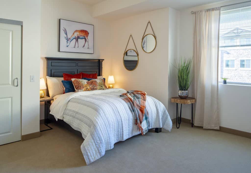 A cozy bedroom with a striped bedspread, decorative pillows, wall art of a deer, and hanging mirrors, complemented by soft lighting and a potted plant.