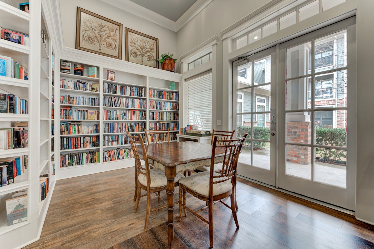 A cozy library with a large bookshelf, a wooden table and chairs, and ample natural light from French doors.