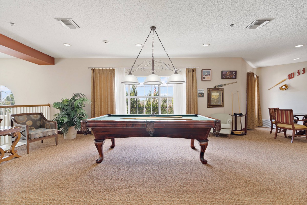 A well-lit game room features a classic pool table centered under a hanging light fixture, with a large window and decorative elements in the background.