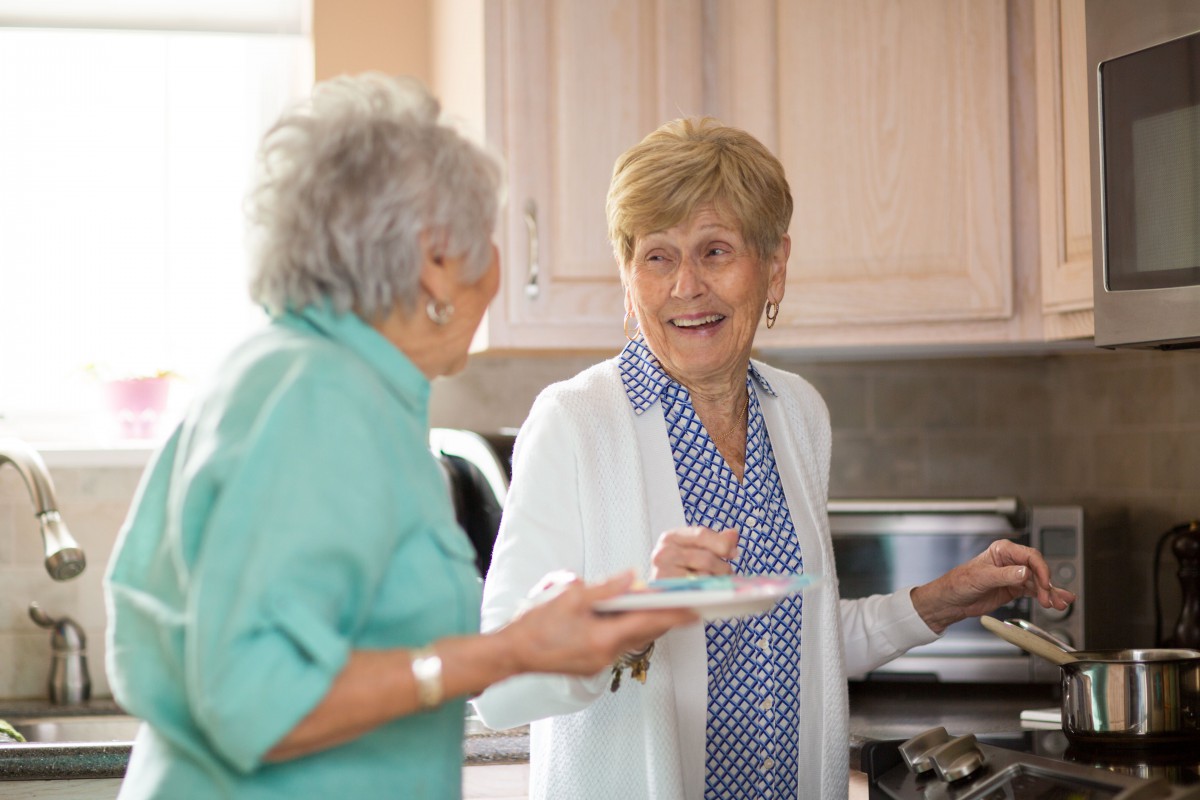 Two elderly women smiling and conversing in the kitchen.