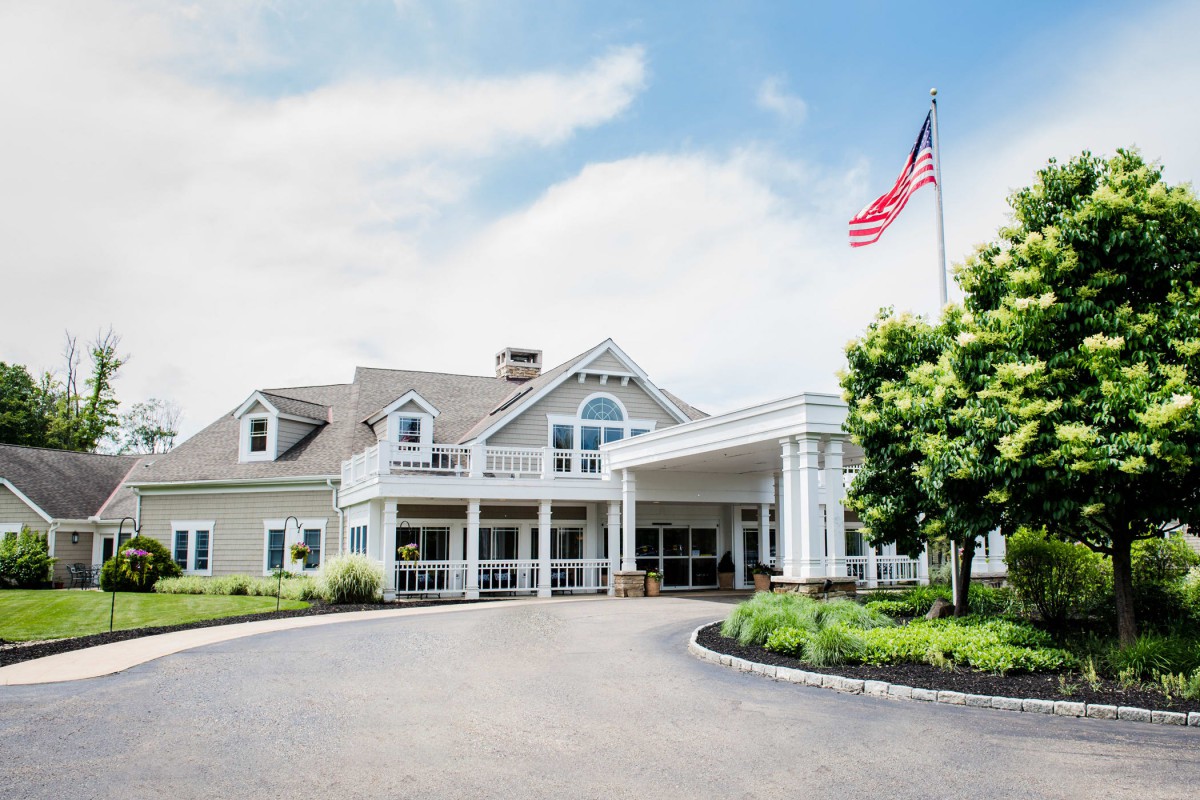 A large building with a sloping roof and a porch area, surrounded by greenery and an American flag flying on the right side.