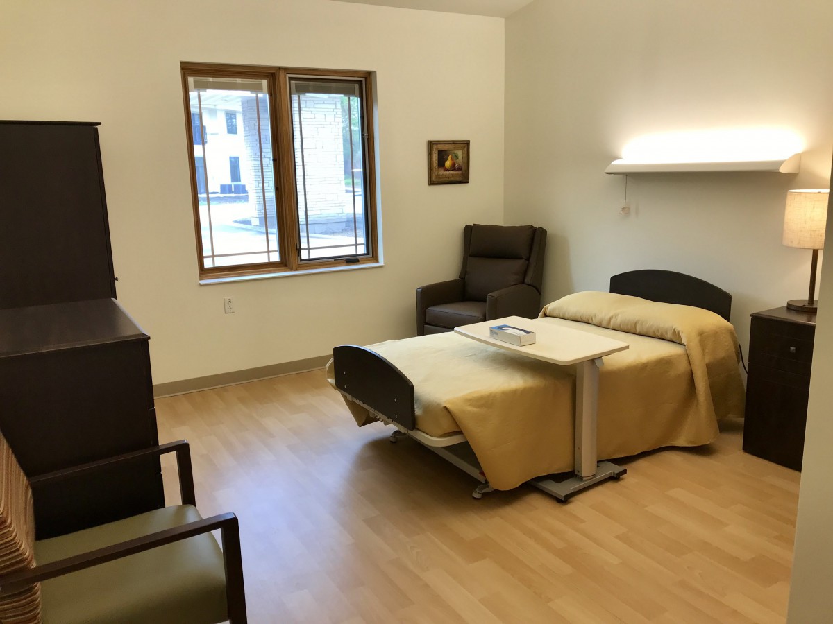 A neatly arranged hospital room with a single bed, chairs, and a window allowing natural light.