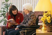 A caregiver reads to an elderly woman sitting in a wheelchair indoors.