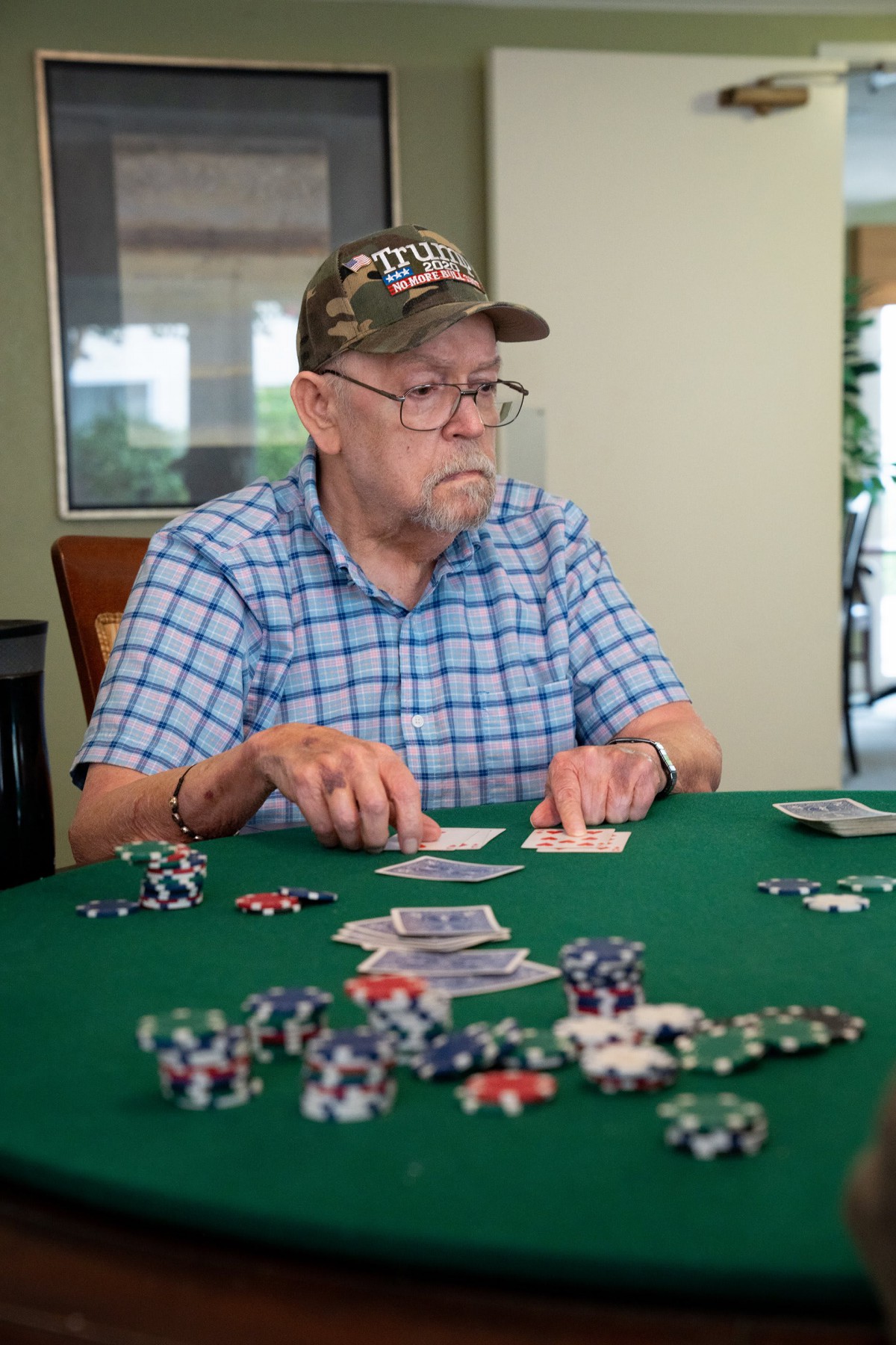 An elderly man wearing a hat plays poker at a table full of chips and cards.