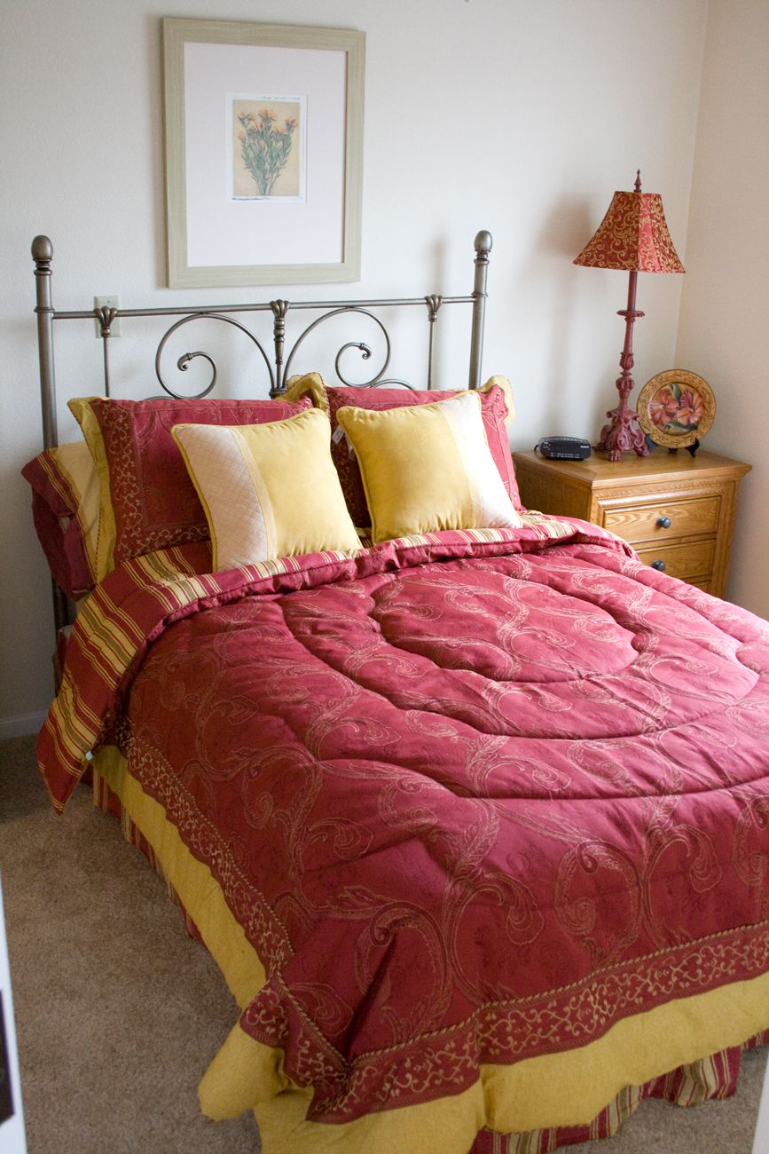 A cozy bedroom with a neatly made bed featuring red and yellow bedding and pillows, accompanied by a nightstand and lamp.