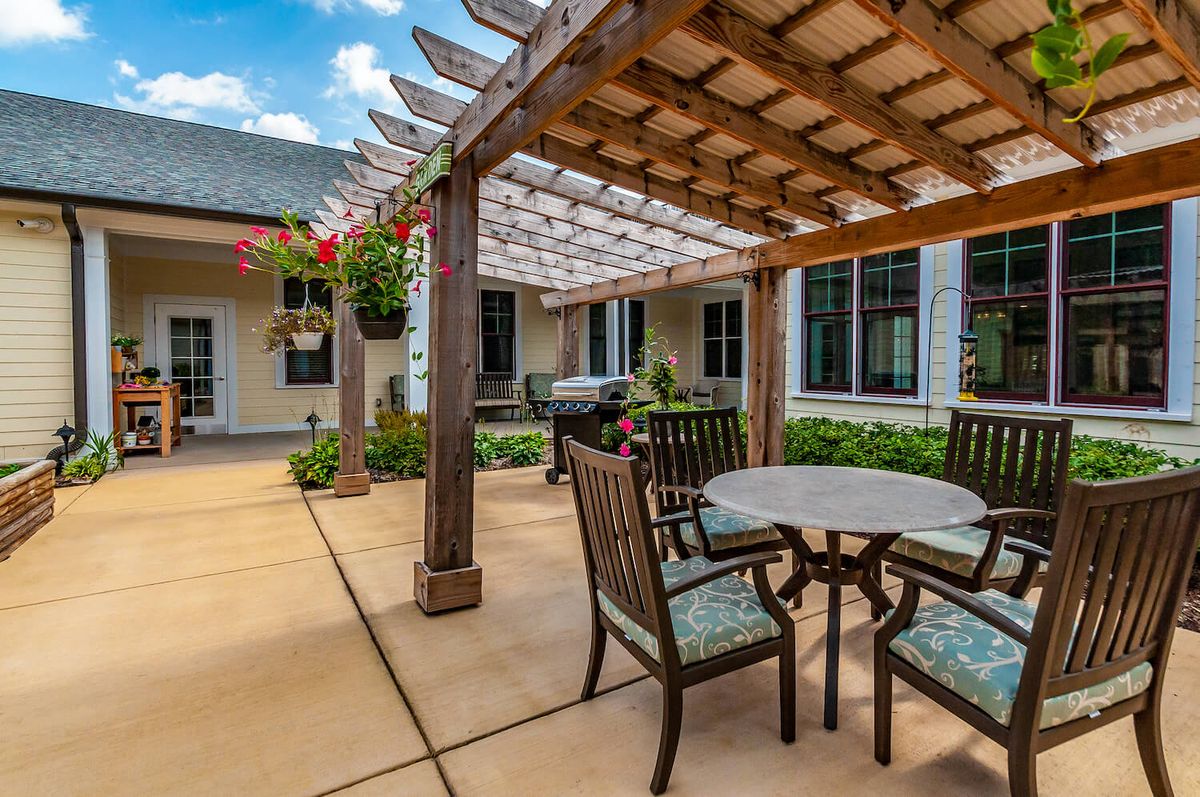 A cozy outdoor patio with a wooden pergola, surrounded by greenery and furnished with a table and chairs.