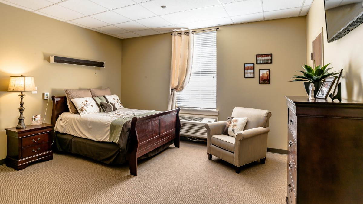 A cozy bedroom with a bed, an armchair, and a window, featuring soft lighting and simple decor.