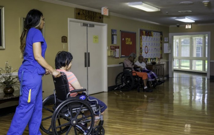 A caregiver assists an elderly person in a wheelchair down the hallway of a nursing home.