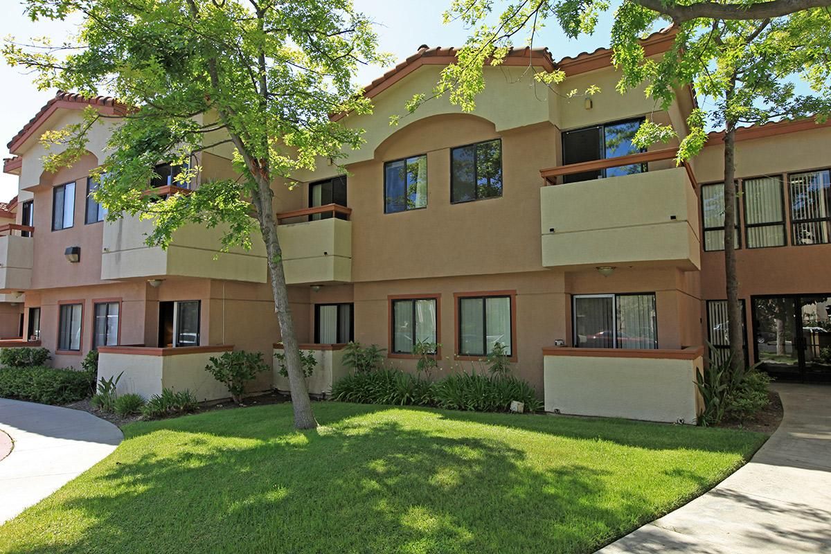 A two-story apartment building with balconies, surrounded by greenery on a sunny day.