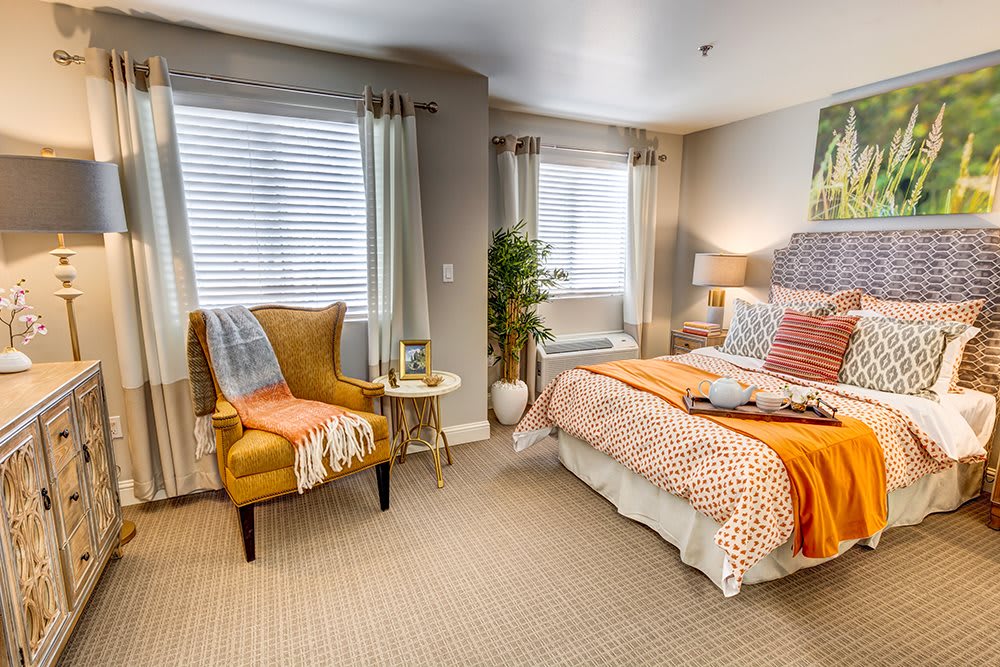 A cozy and modern bedroom with orange accents, featuring a comfortable bed, armchair, and ample natural light.