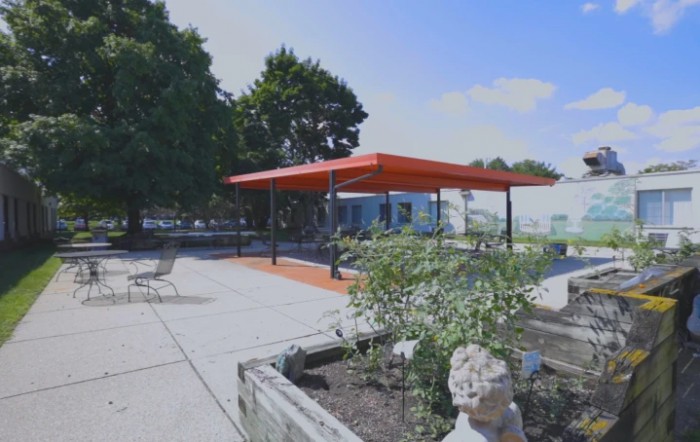 A garden patio area with a red canopy, surrounded by greenery and sculptures.