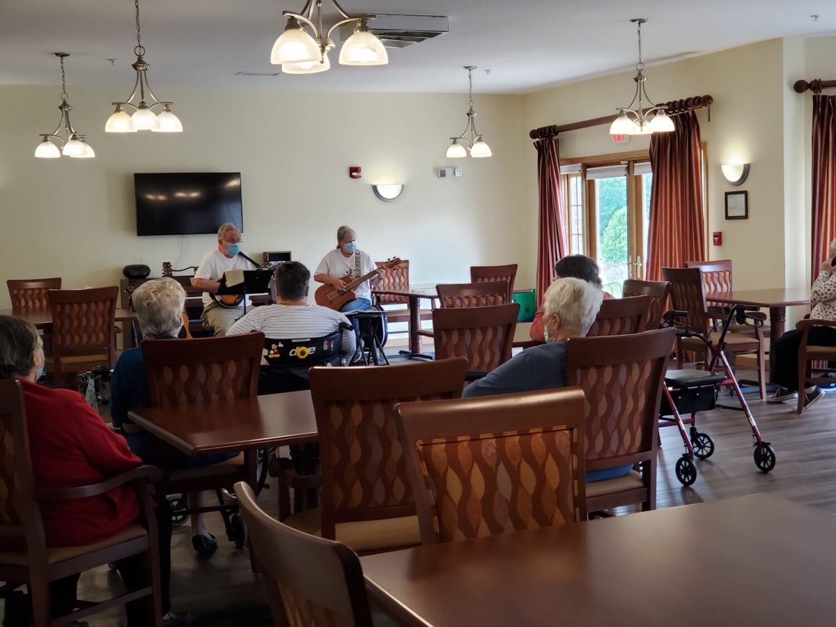 A small gathering of people watching musicians perform in a senior home common area.