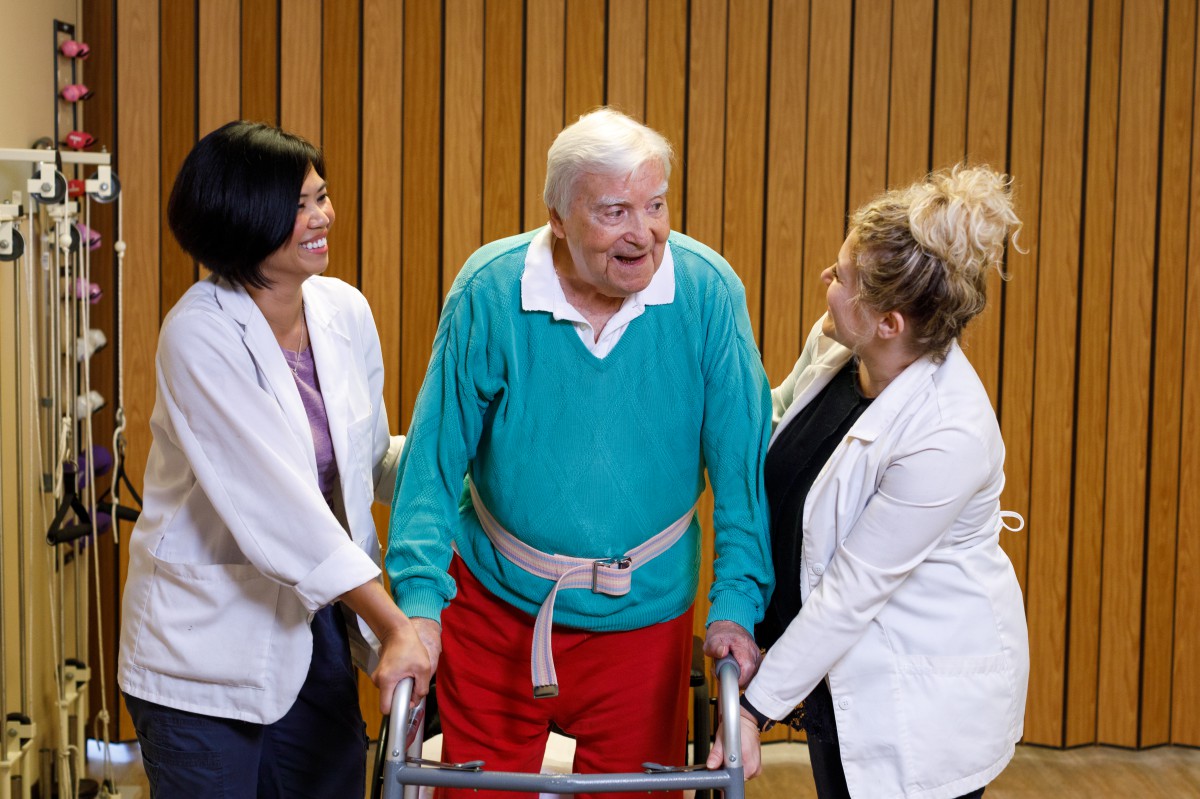 An elderly man using a walker is assisted by two healthcare professionals.