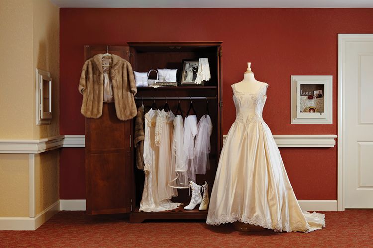 A vintage wedding dress displayed on a mannequin next to an open wardrobe containing elegant garments, a fur coat, and white boots against a red wall.