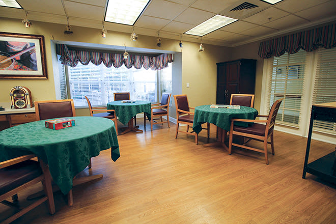 A well-lit dining area with round tables and chairs, featuring large windows and green tablecloths.