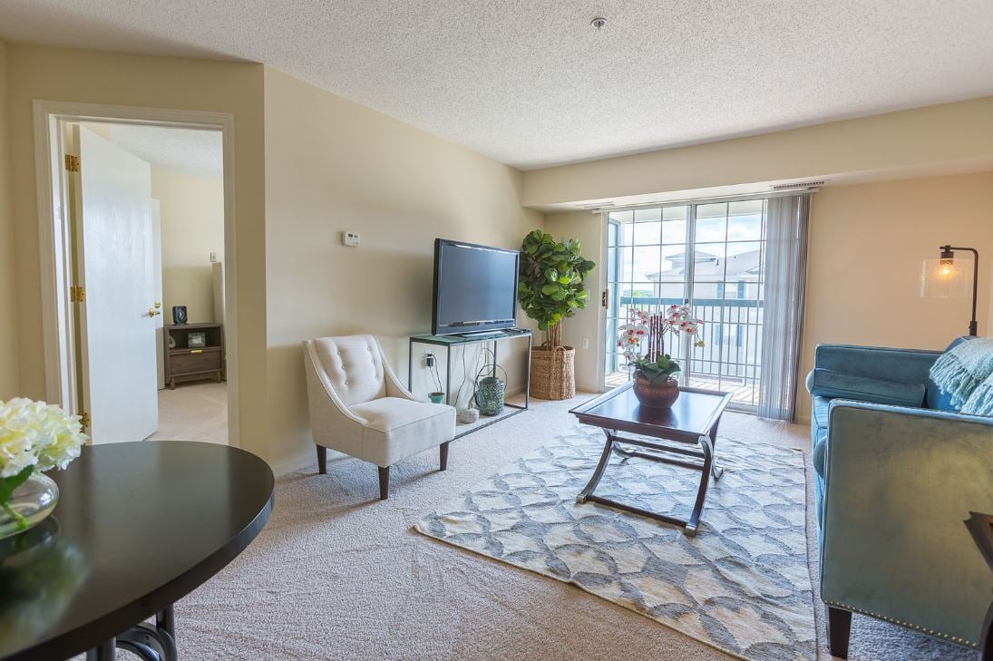 A well-lit living room featuring comfortable seating, a television, and a coffee table, with large windows letting in natural light.