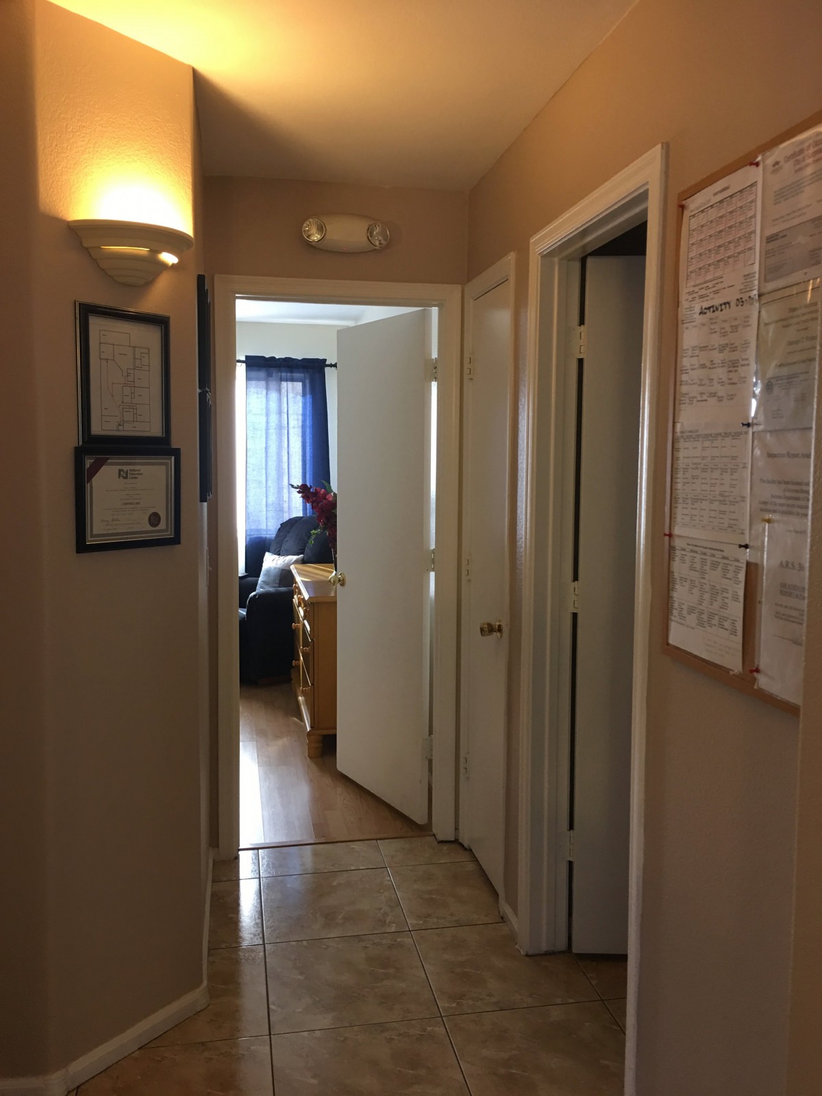 A well-lit hallway with open doors leading to a room, featuring wall-mounted documents and framed certificates.