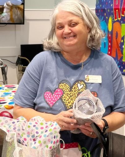 An elderly woman smiling and holding a gift at a celebration indoors.