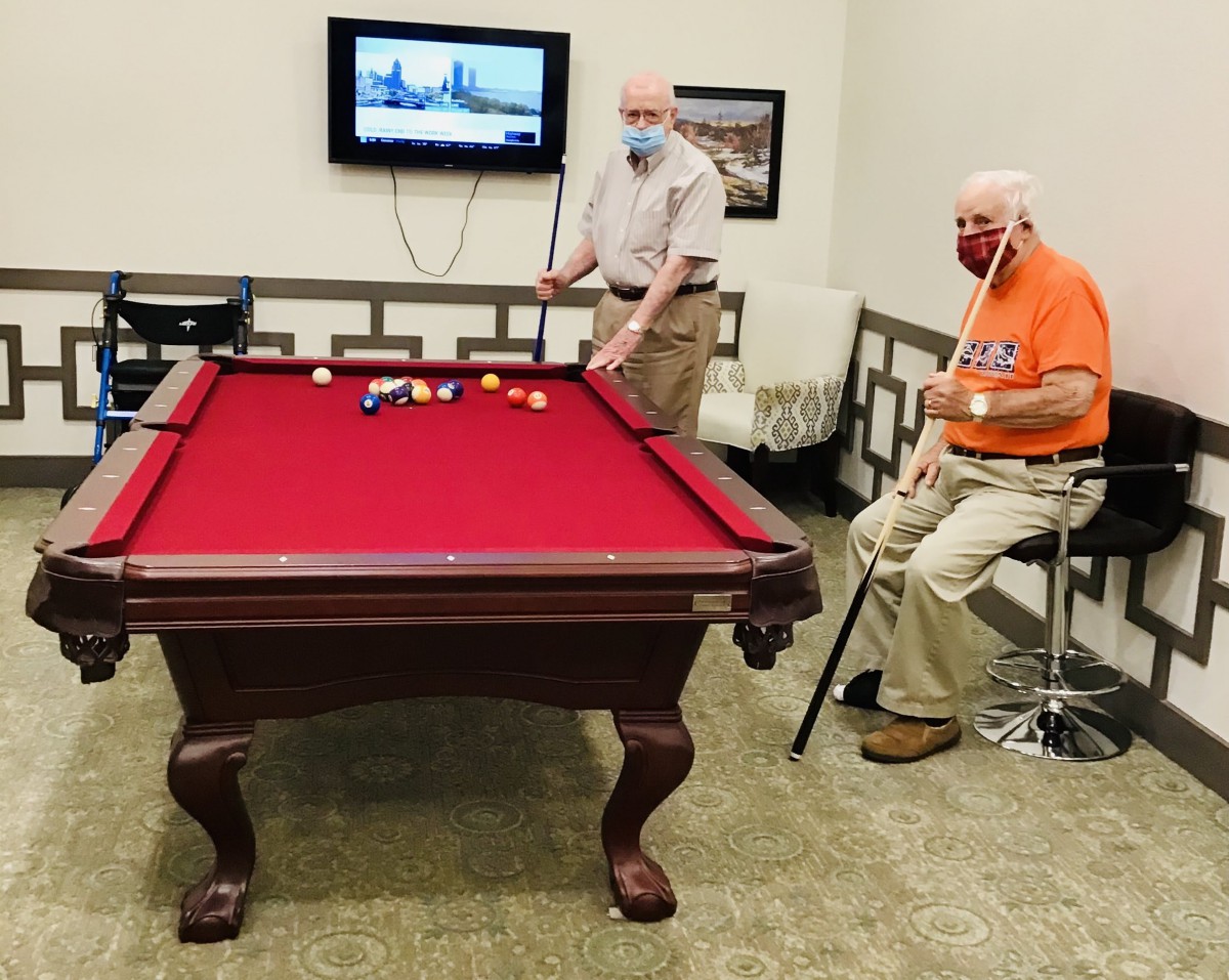 Two elderly individuals playing pool indoors while wearing masks.