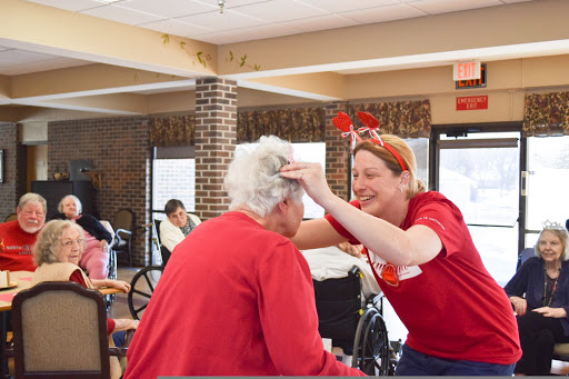 A caregiver joyfully interacts with an elderly resident in a community setting, both wearing red to celebrate a special occasion.