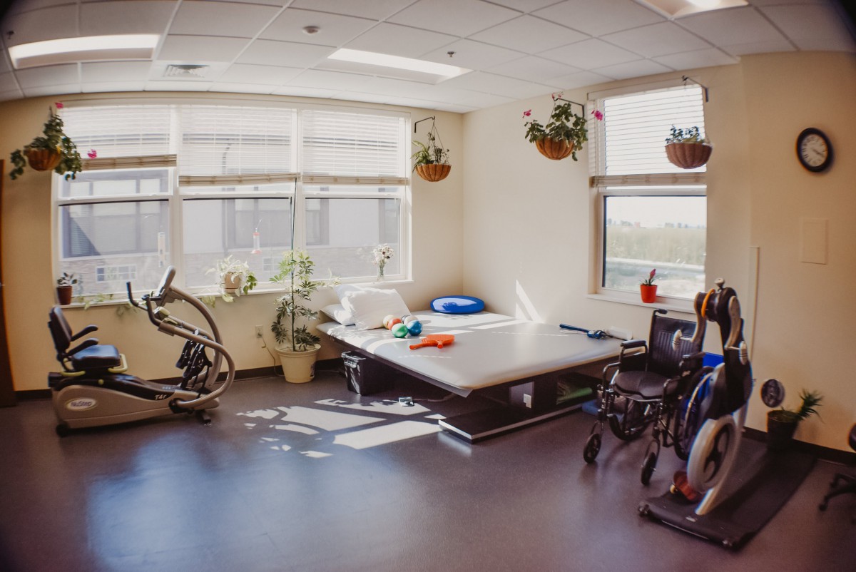 A well-lit rehabilitation room equipped with exercise equipment and a wheelchair, decorated with hanging plants.