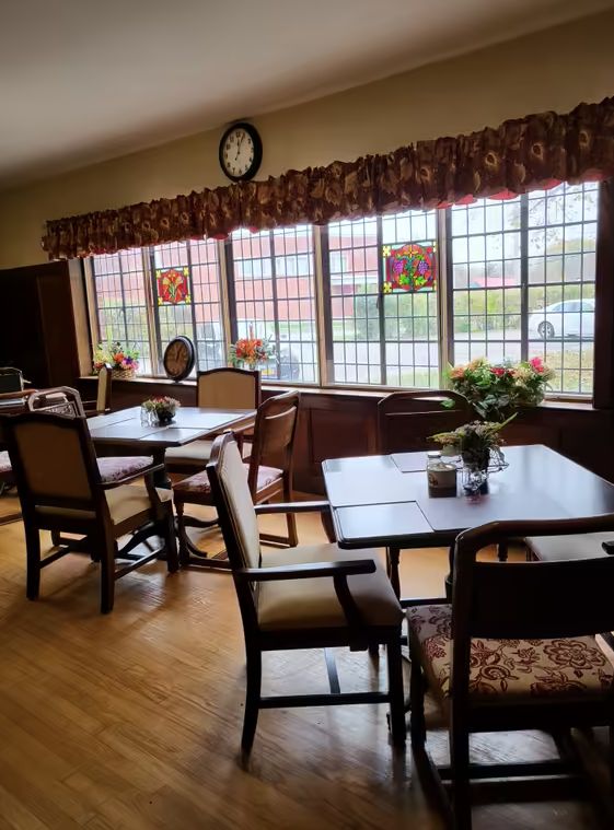 A cozy dining area with neatly arranged tables and chairs, decorated with flowers and illuminated by natural light through large windows.
