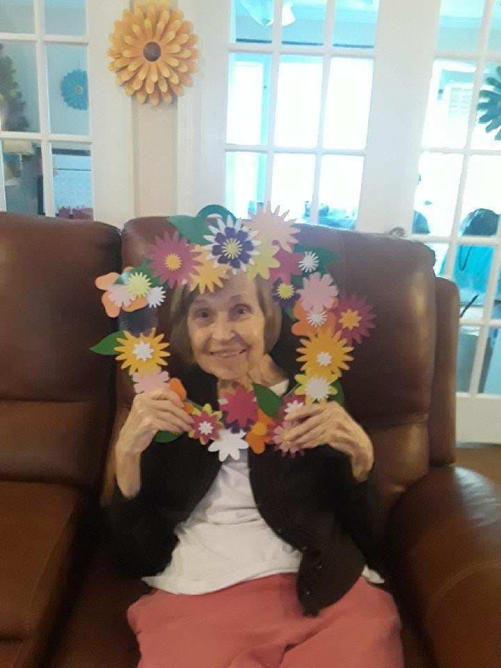 An elderly woman smiling while holding a colorful flower frame in a living room.