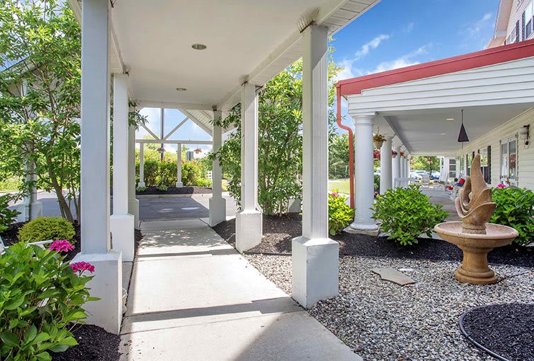 A covered walkway with columns leading to an entrance, surrounded by landscaped gardens.
