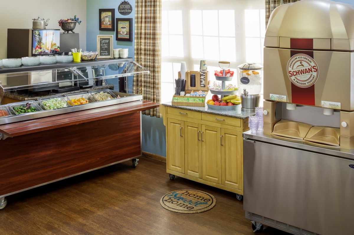 A kitchen interior with a buffet setup, featuring a coffee maker, fruits, and various food displays on cabinets.