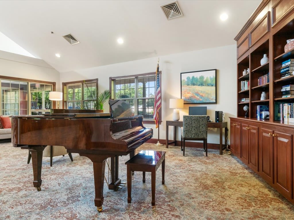 A cozy living room with a grand piano and office area featuring a bookshelf, desk, and artwork.