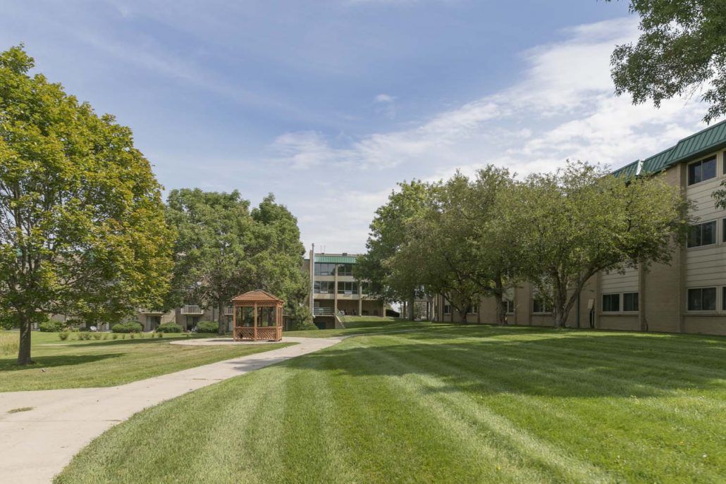 A sunny day on a campus with a well-maintained lawn, pathway, gazebo, and surrounding trees.