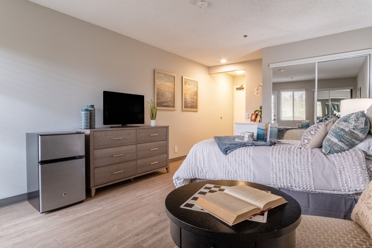 A cozy, modern bedroom with a bed, dresser, and television, featuring warm lighting and an open book on the table.