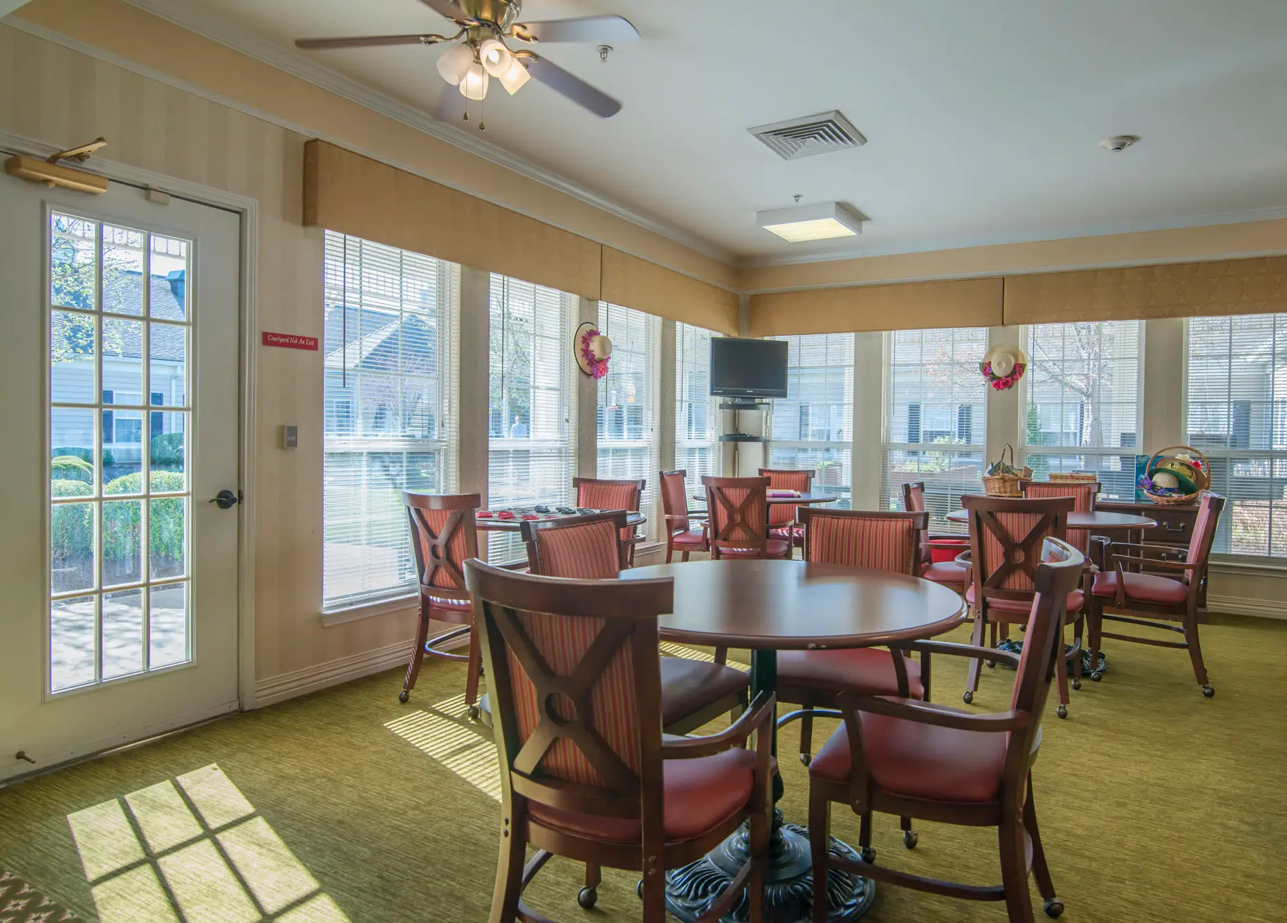 A cozy dining room with multiple tables and chairs, well-lit by large windows and a ceiling fan.
