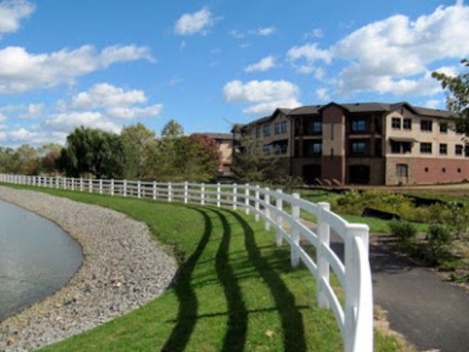 Scenic view of a white fence curving around a pond, with apartment buildings in the background and a blue sky above.