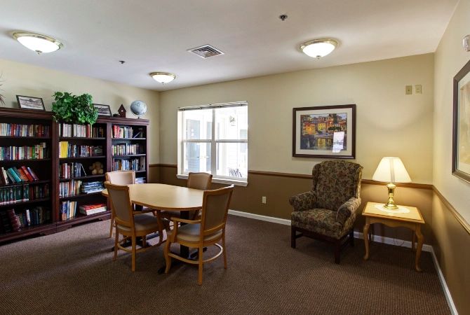 A cozy library room with a table, chairs, and a bookshelf near a window.