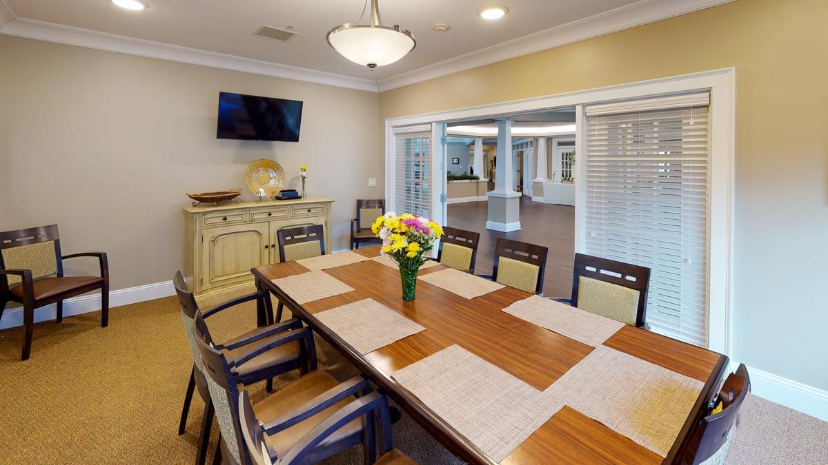A well-lit dining room featuring a wooden table with placemats, surrounded by chairs and a vase of flowers in the center.