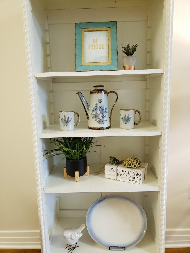 A neatly arranged shelf featuring decorative items including a teapot, mugs, framed art, and plants.