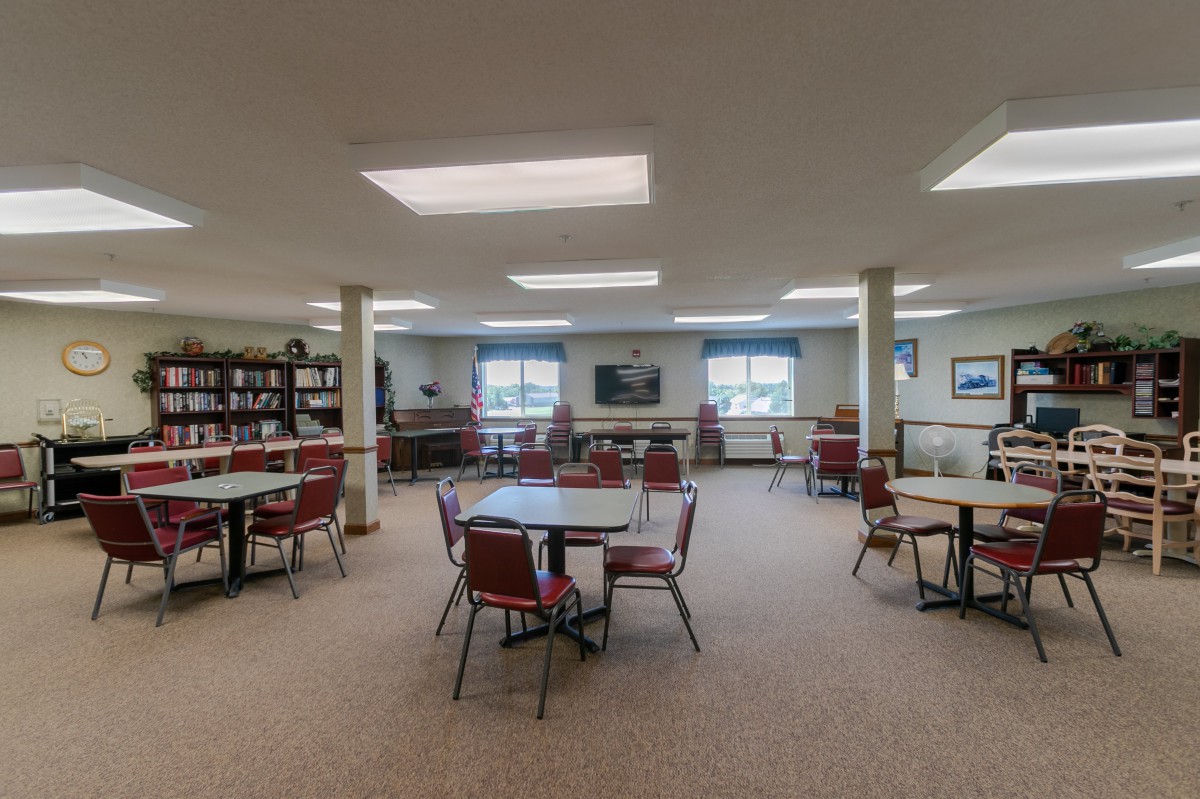 A spacious library interior with tables and chairs, surrounded by bookshelves and well-lit by large windows.