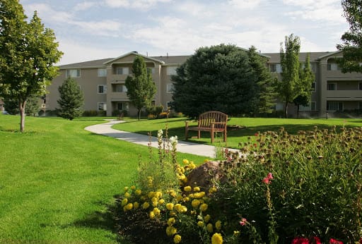 A landscaped garden area with blooming flowers, trees, a bench, and an apartment building in the background.