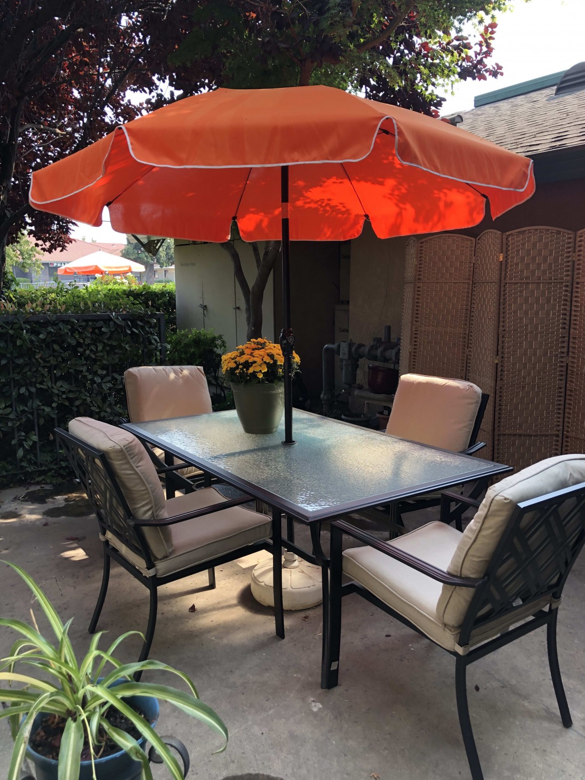 A cozy patio setup with a glass table, cushioned chairs, and an orange umbrella providing shade.