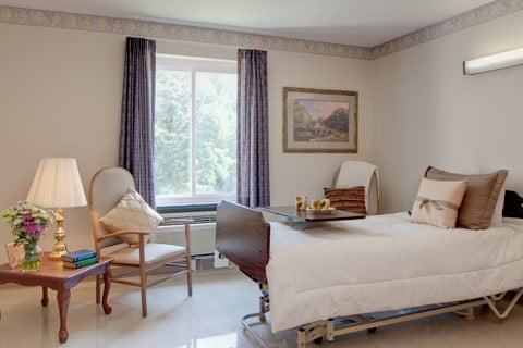 A serene hospital room featuring a bed, comfortable chair, and natural light streaming through the window.